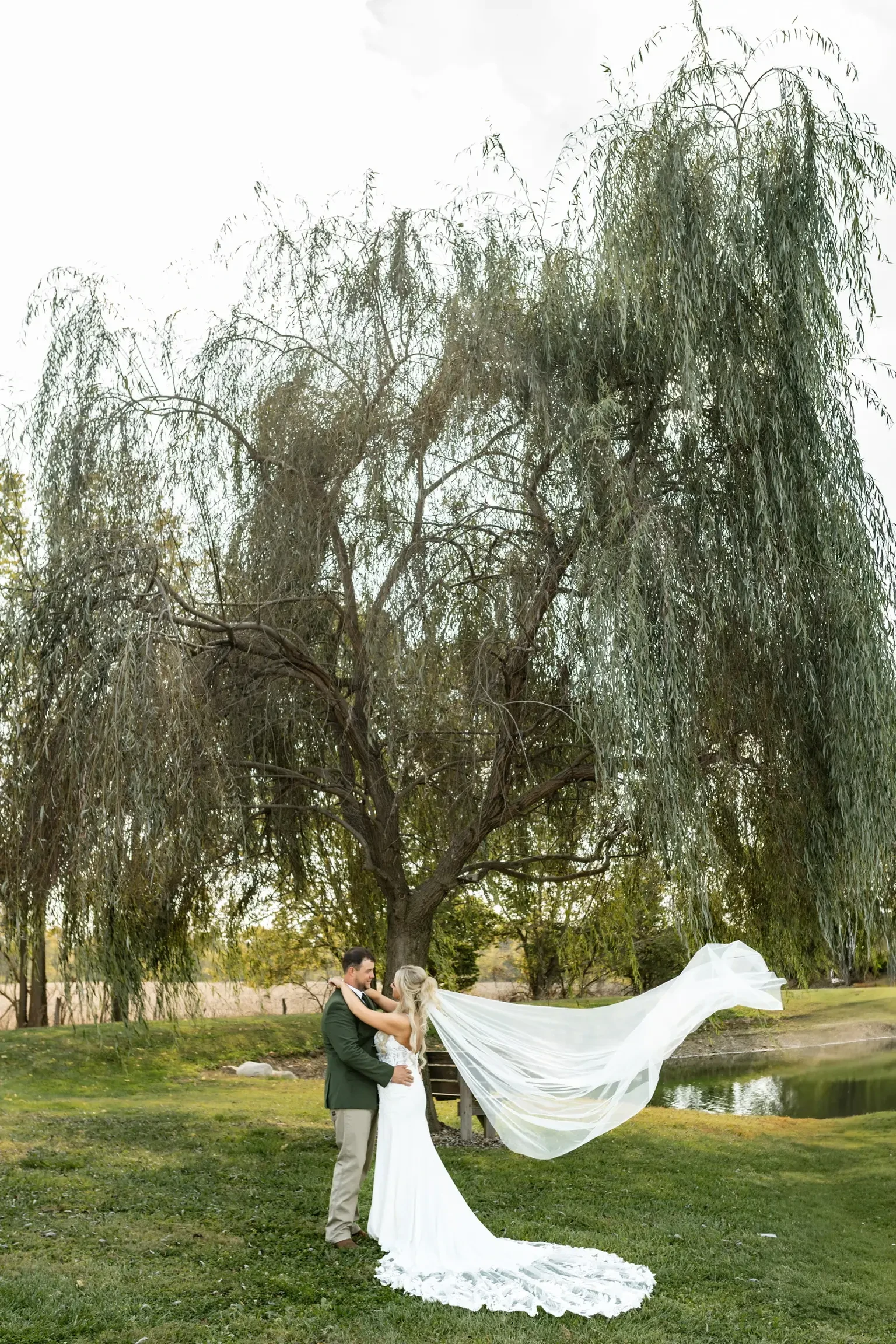 A bride and groom embrace under a large willow tree by a pond. The bride's veil flows gracefully in the breeze, creating a serene, romantic scene.
