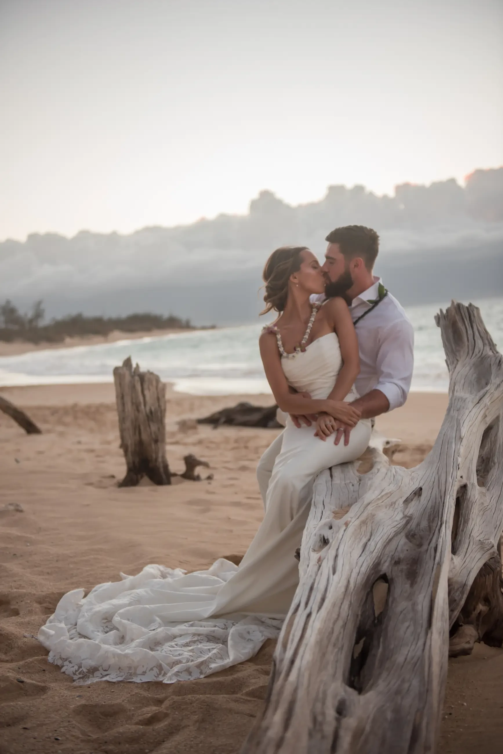 A couple in wedding attire sharing a kiss on a beach, sitting on a piece of driftwood.