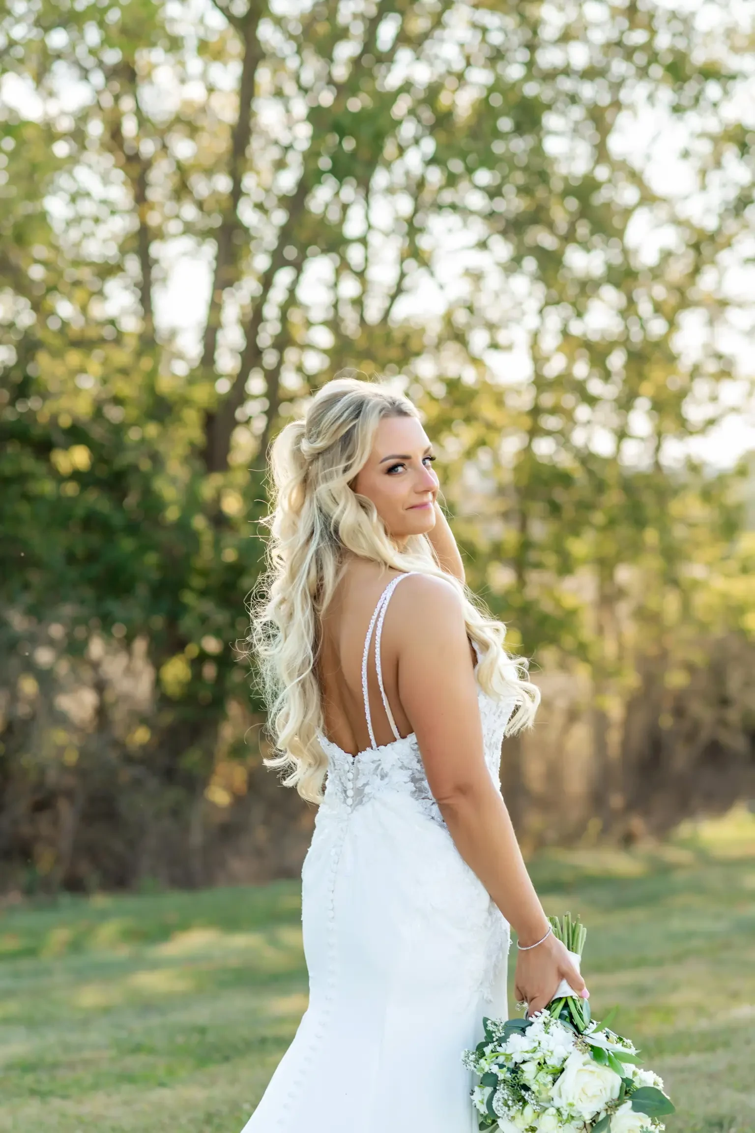 Bride with long blonde curls in a white gown holds a bouquet, gazing back with a smile. She stands outside, surrounded by sunlit trees and green grass.