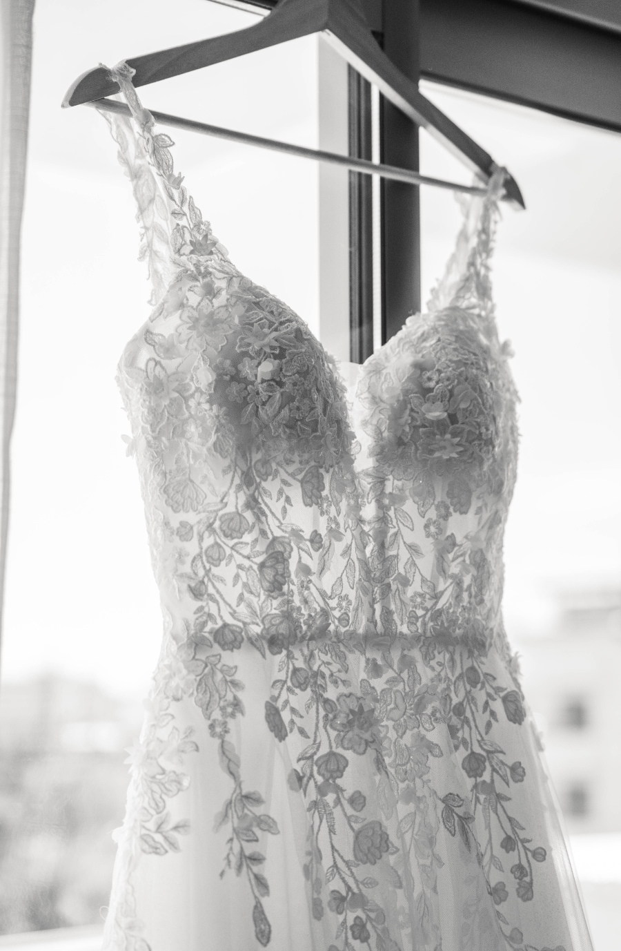Elegant lace wedding dress on hanger by a window. Sunlight highlights floral patterns, creating a romantic, serene atmosphere.
