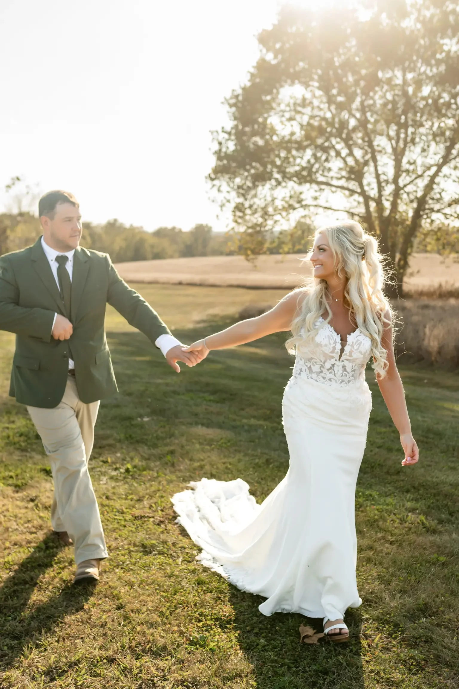 A couple holds hands, walking on a grassy field. The woman in a white bridal gown gazes at her partner, who wears a green jacket. Sunlight filters through trees.