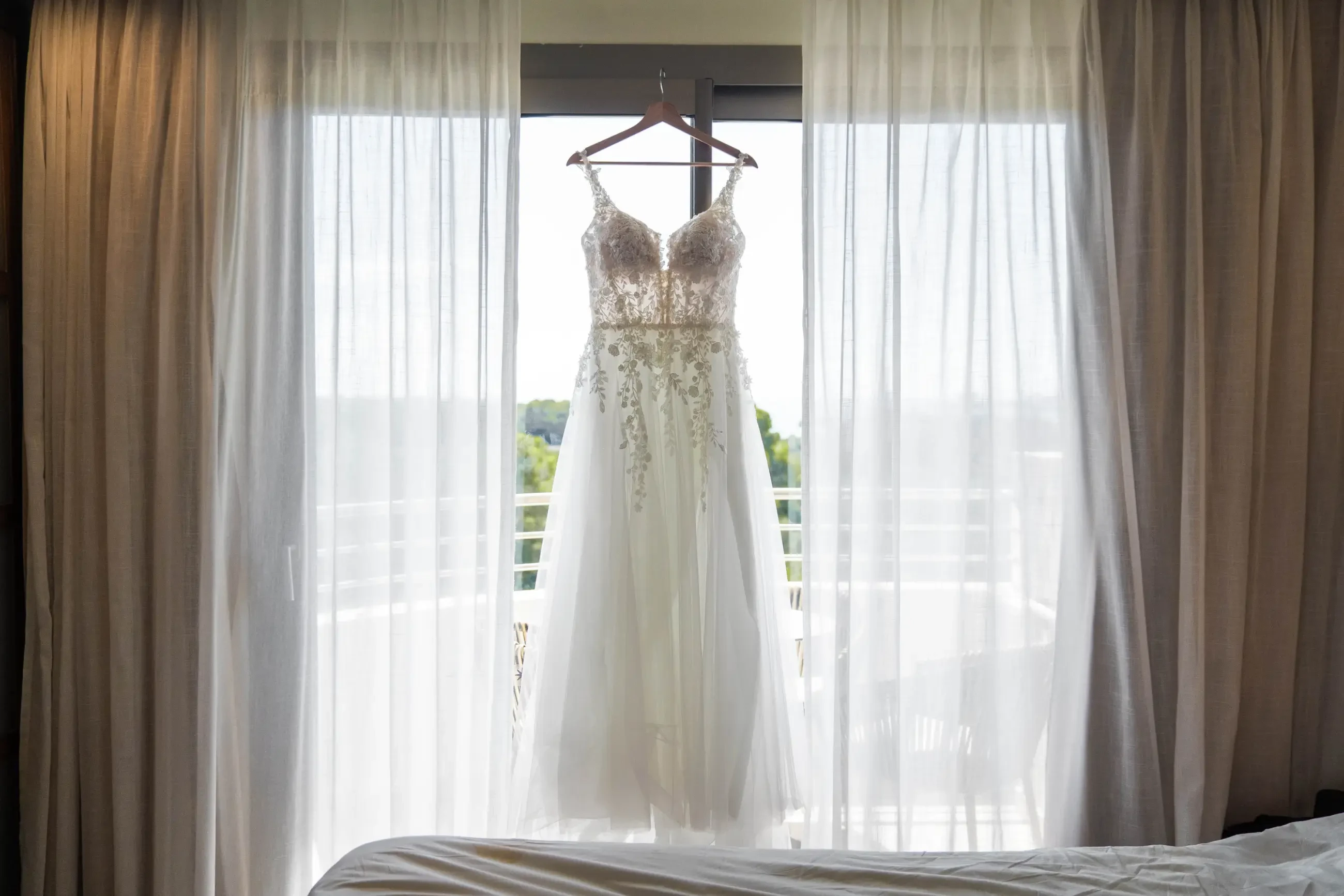 A delicate white wedding dress with intricate lace details hangs in front of sheer curtains, backlit by soft natural light, creating a serene, elegant ambiance.