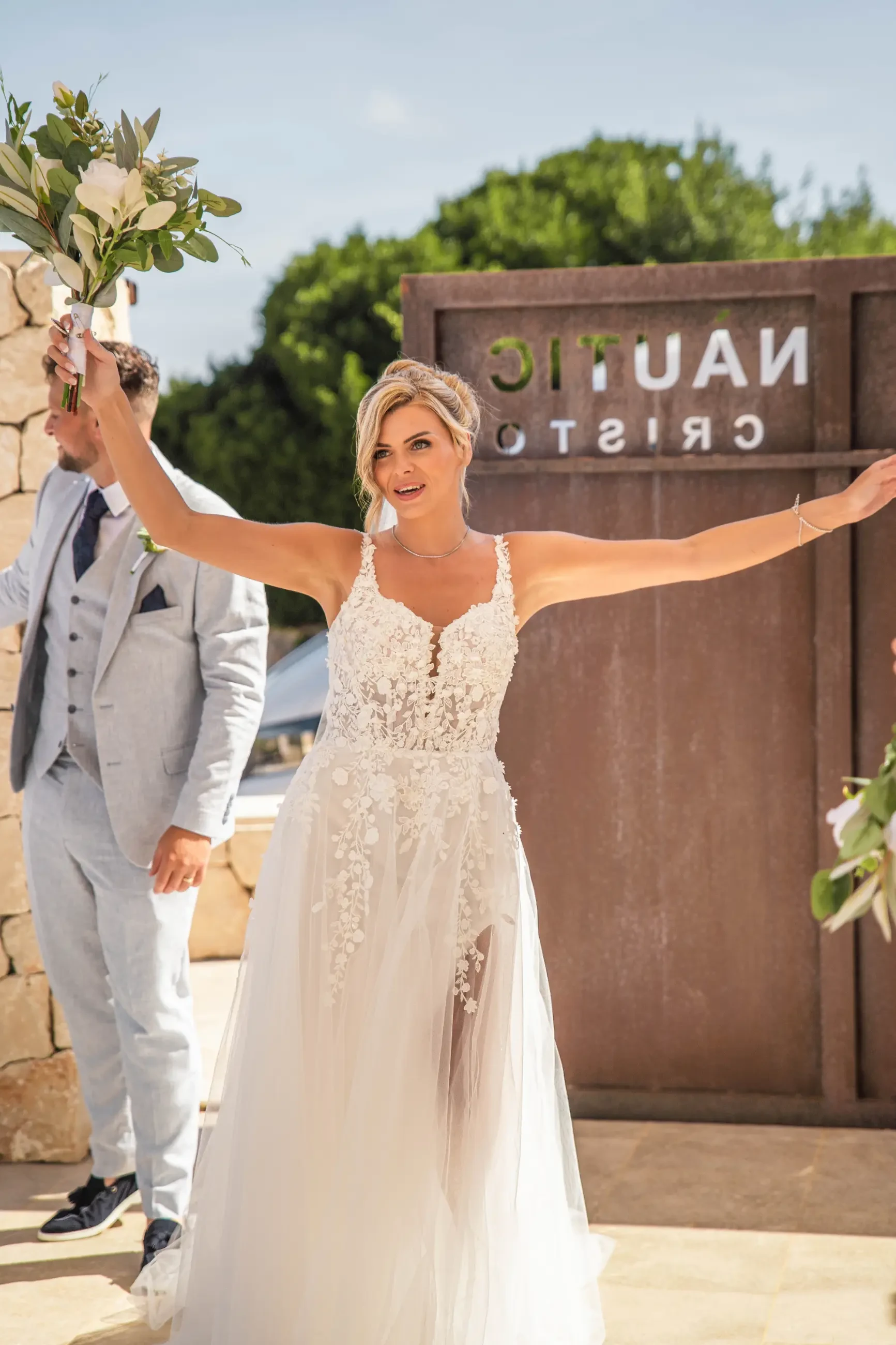 A joyful bride in a white lace gown holds a bouquet high, celebrating. A man in a gray suit stands beside her. Background shows green trees and a rusted sign.
