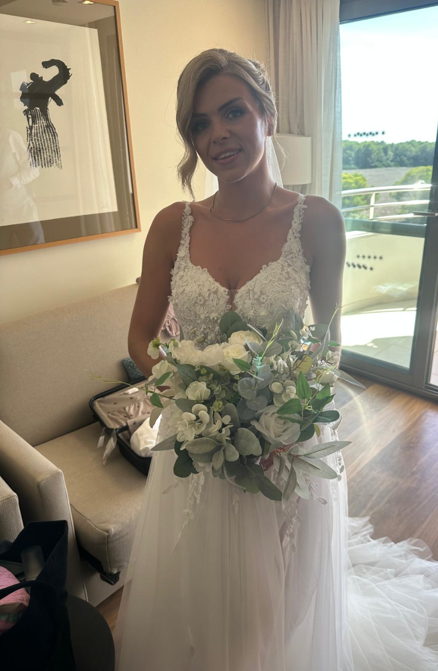 Bride in lace wedding dress, holding a white and green bouquet, standing in a bright room near a window with a view of trees. Calm and joyful atmosphere.