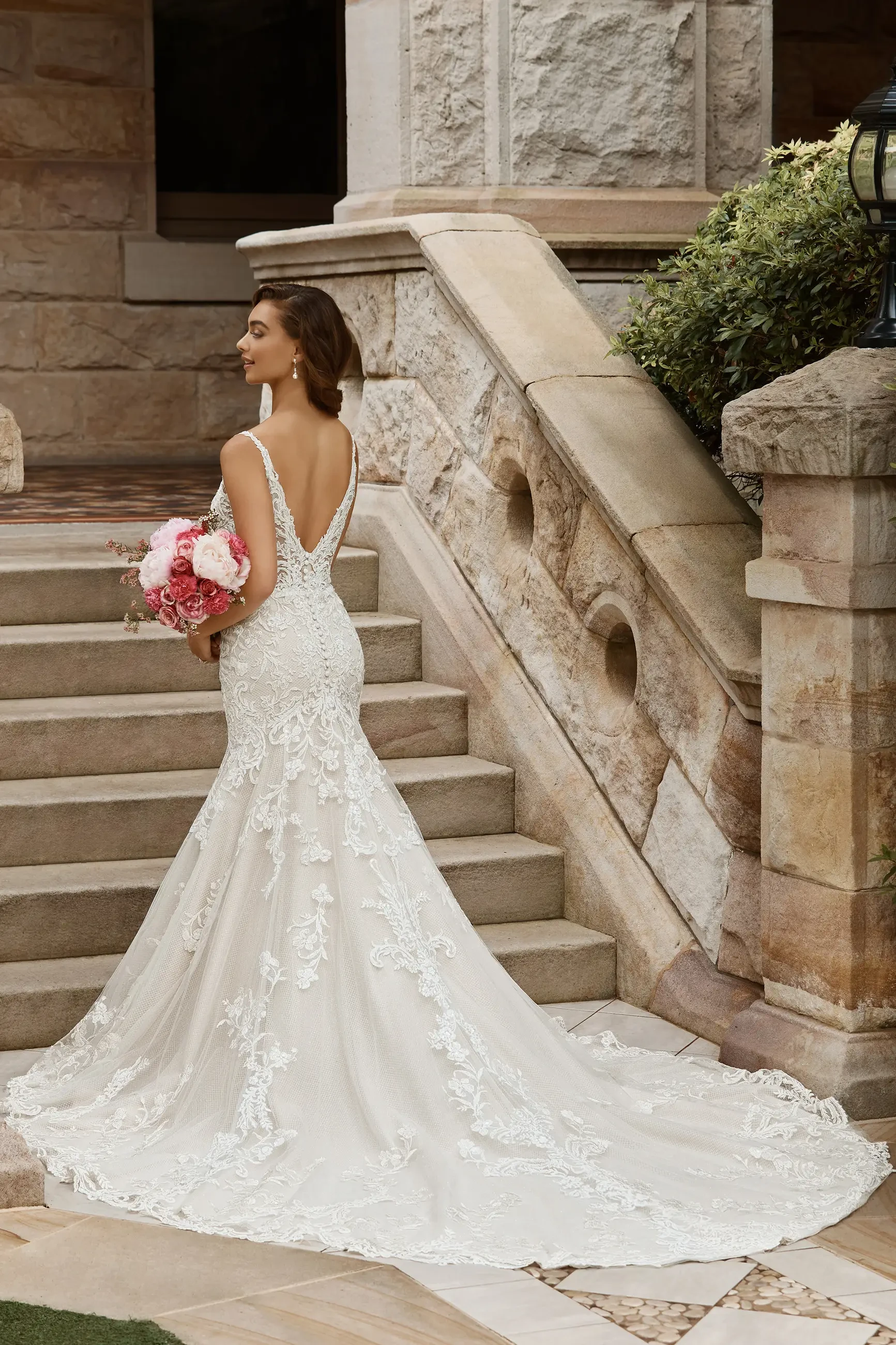 A woman in an elegant wedding dress stands on steps, holding a bouquet of pink flowers. The dress features lace details and a long train, and she is facing away from the camera, looking back over her shoulder. The setting is a stone staircase with greenery in the background.