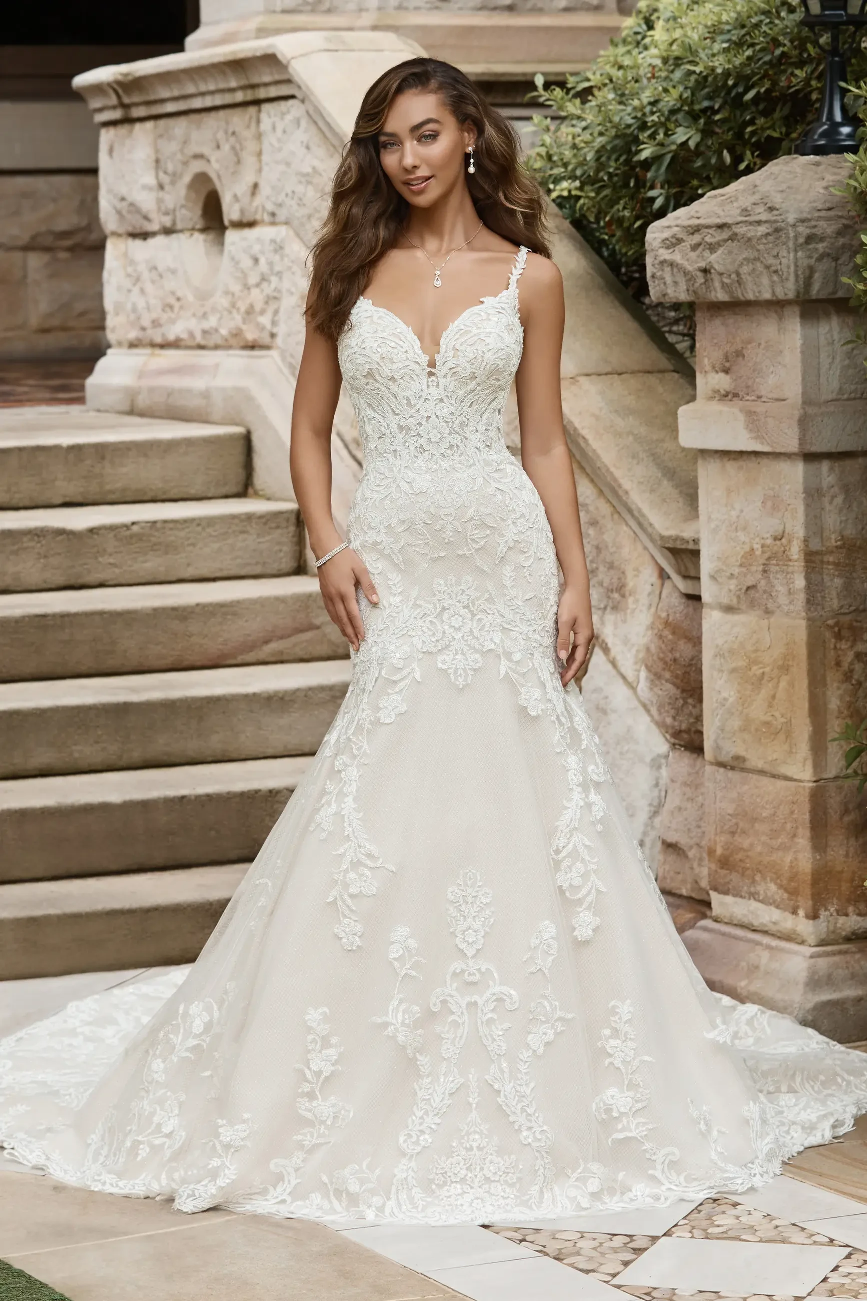 A woman in an elegant white wedding gown with intricate lace details, standing on outdoor steps.