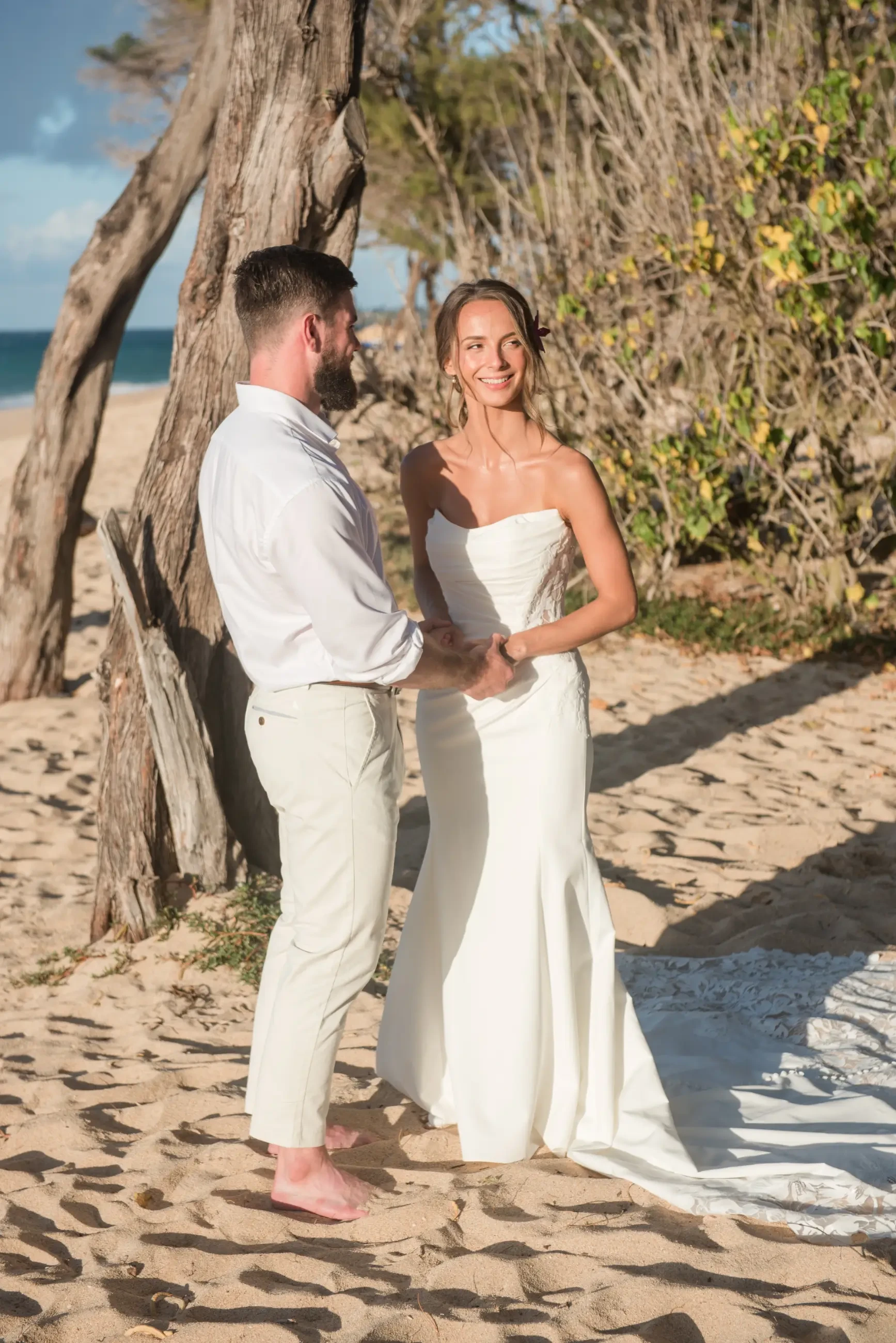 A couple stands barefoot on the beach, holding hands. The woman wears a strapless white wedding gown, and the man is in a white shirt and beige pants. They are smiling at each other with a natural background of sand and greenery.