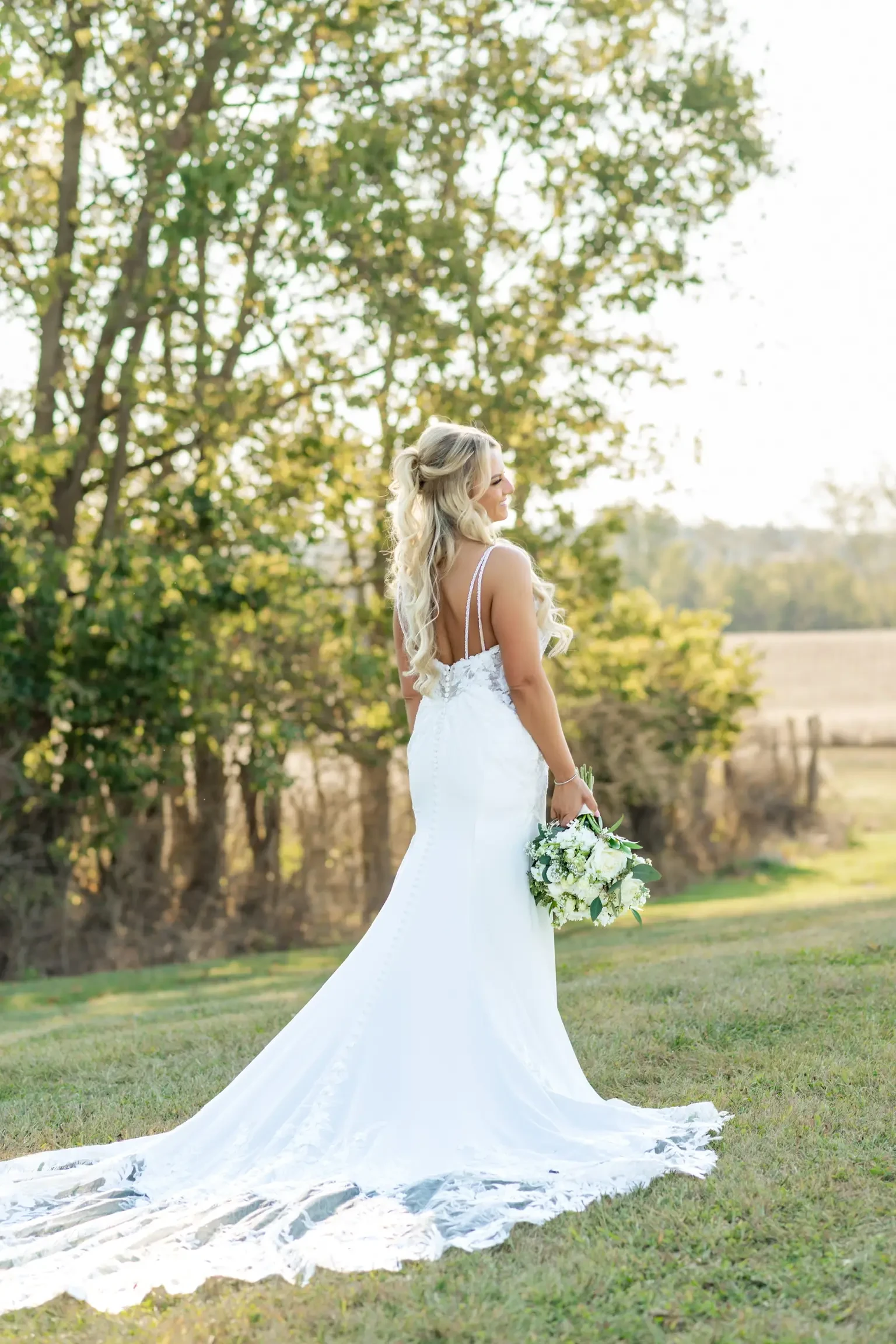 A bride in a flowing white gown stands on grass, holding a bouquet. Sunlight filters through trees in the background, creating a serene, romantic scene.