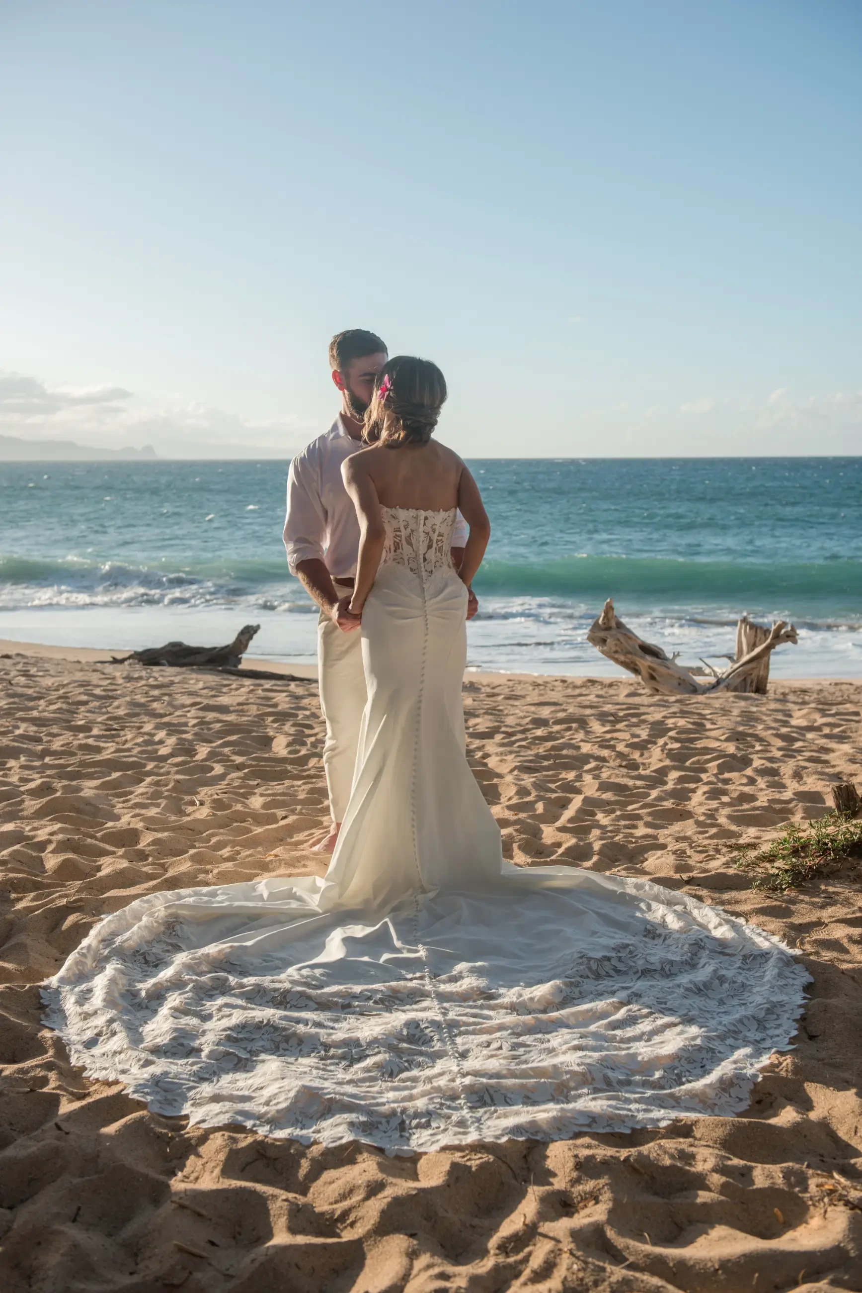 A couple stands on a beach during sunset, gazing at each other. The woman is wearing a white wedding gown, and the man is in light-colored attire. The ocean waves are visible in the background.