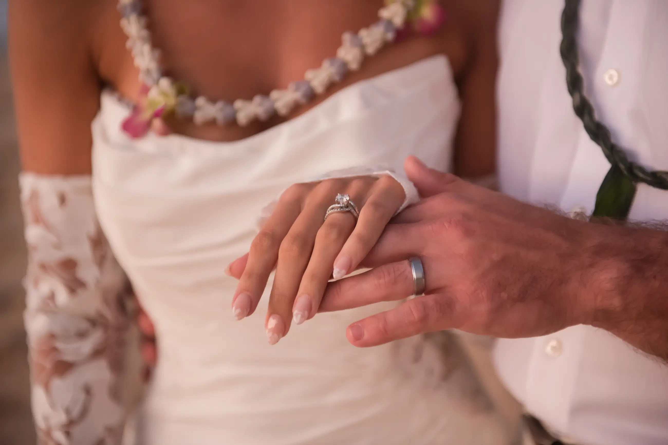 A close-up of a bride and groom's hands, showcasing their wedding rings. The bride&rsquo;s hand is adorned with a diamond ring, while the groom wears a simple band. The couple appears to be in a romantic setting.