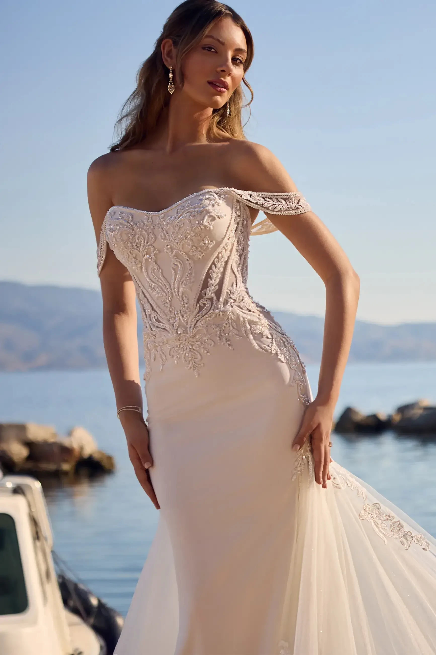 A woman in an elegant off-the-shoulder wedding dress poses by the water with a scenic backdrop.