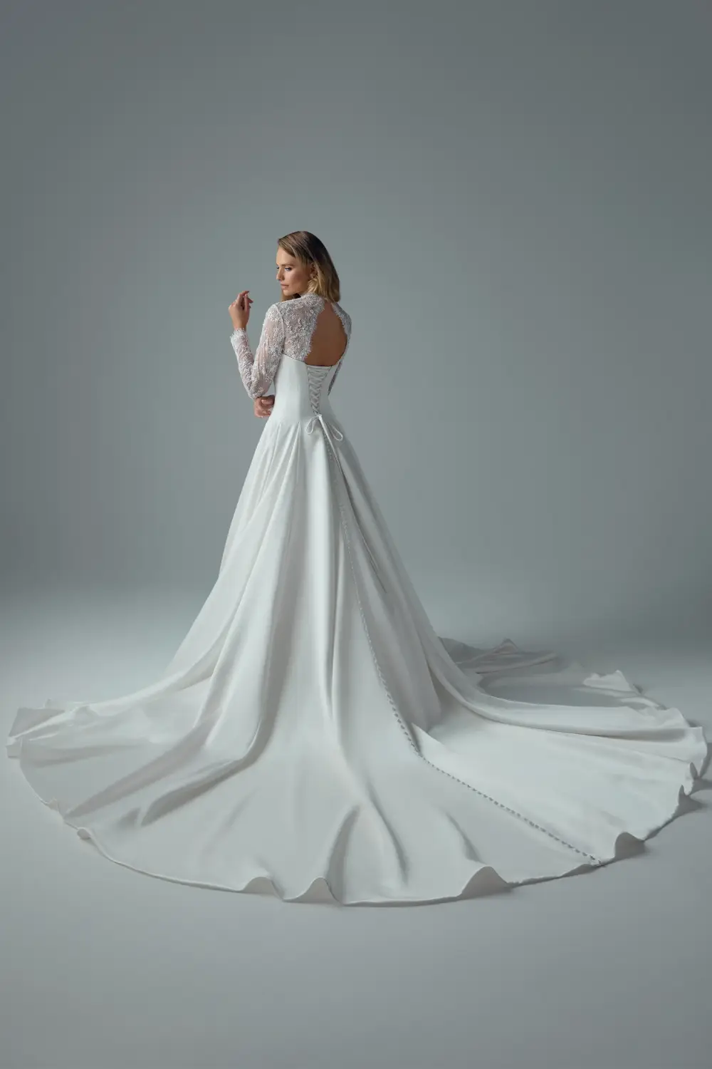 A woman in a white wedding dress with long lace sleeves stands gracefully against a neutral background.