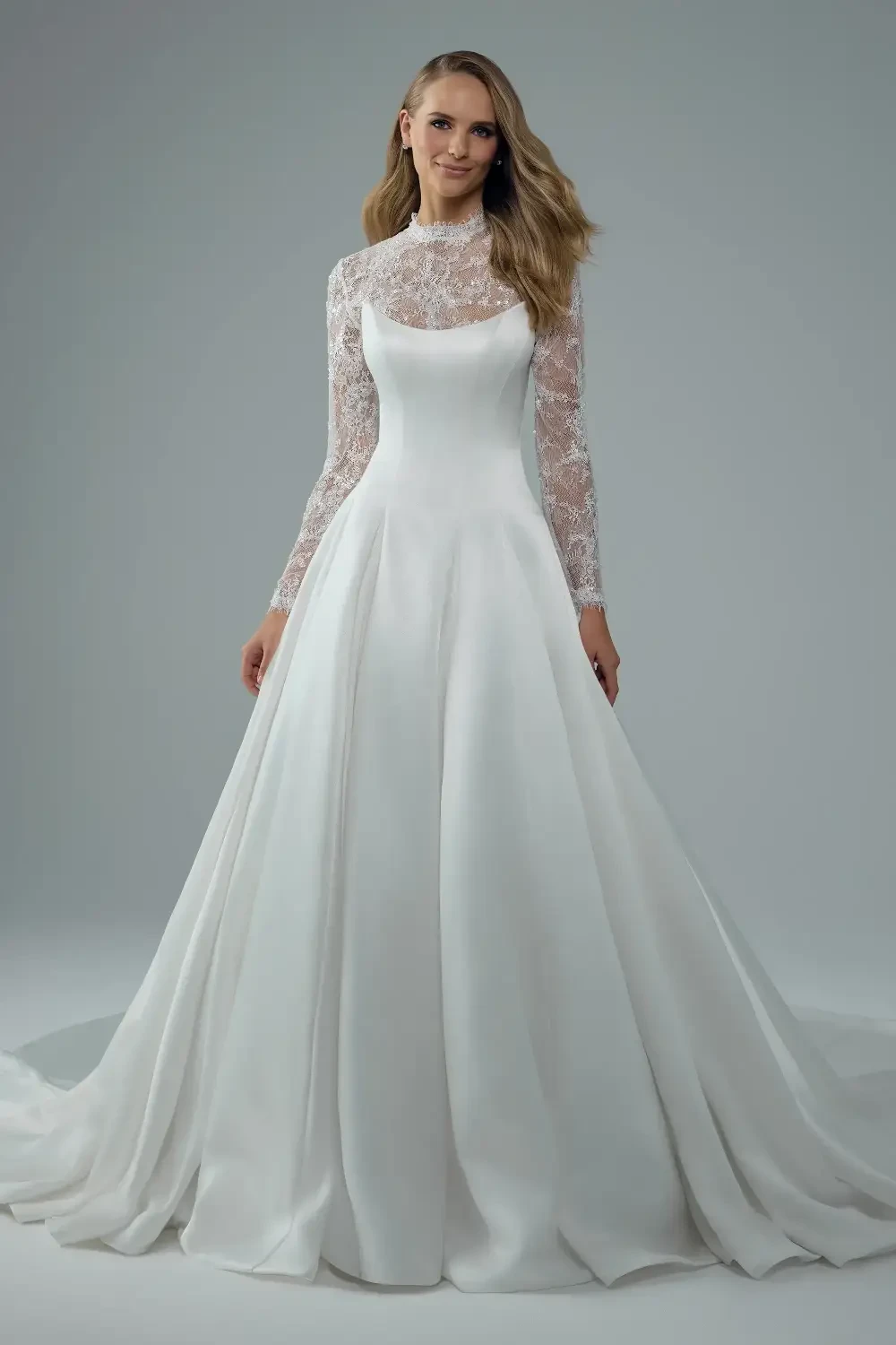 A woman in a white wedding dress with long lace sleeves stands gracefully against a neutral background.