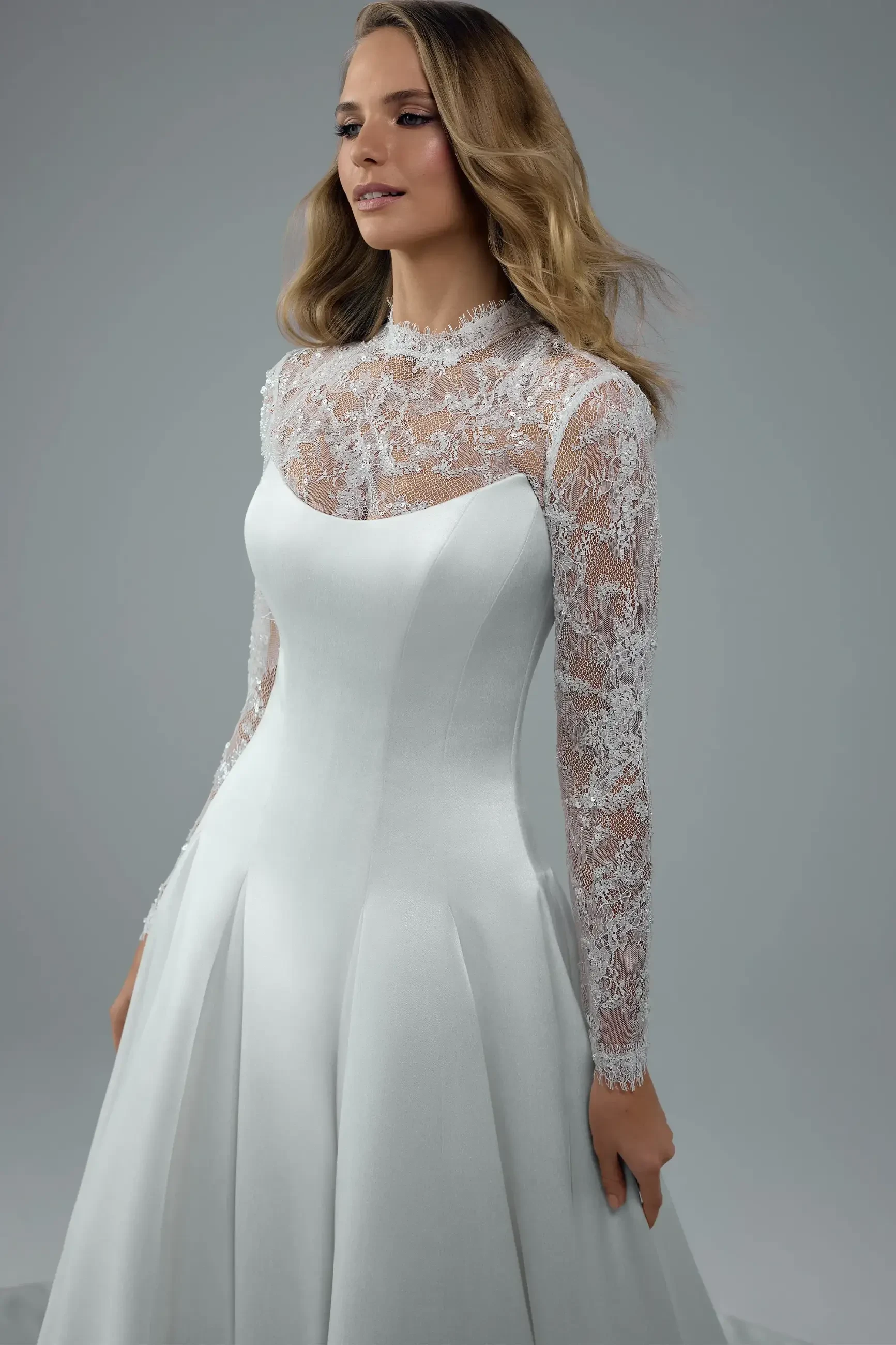 A woman in a white bridal gown with long, sheer lace sleeves and a fitted bodice, standing against a plain background.