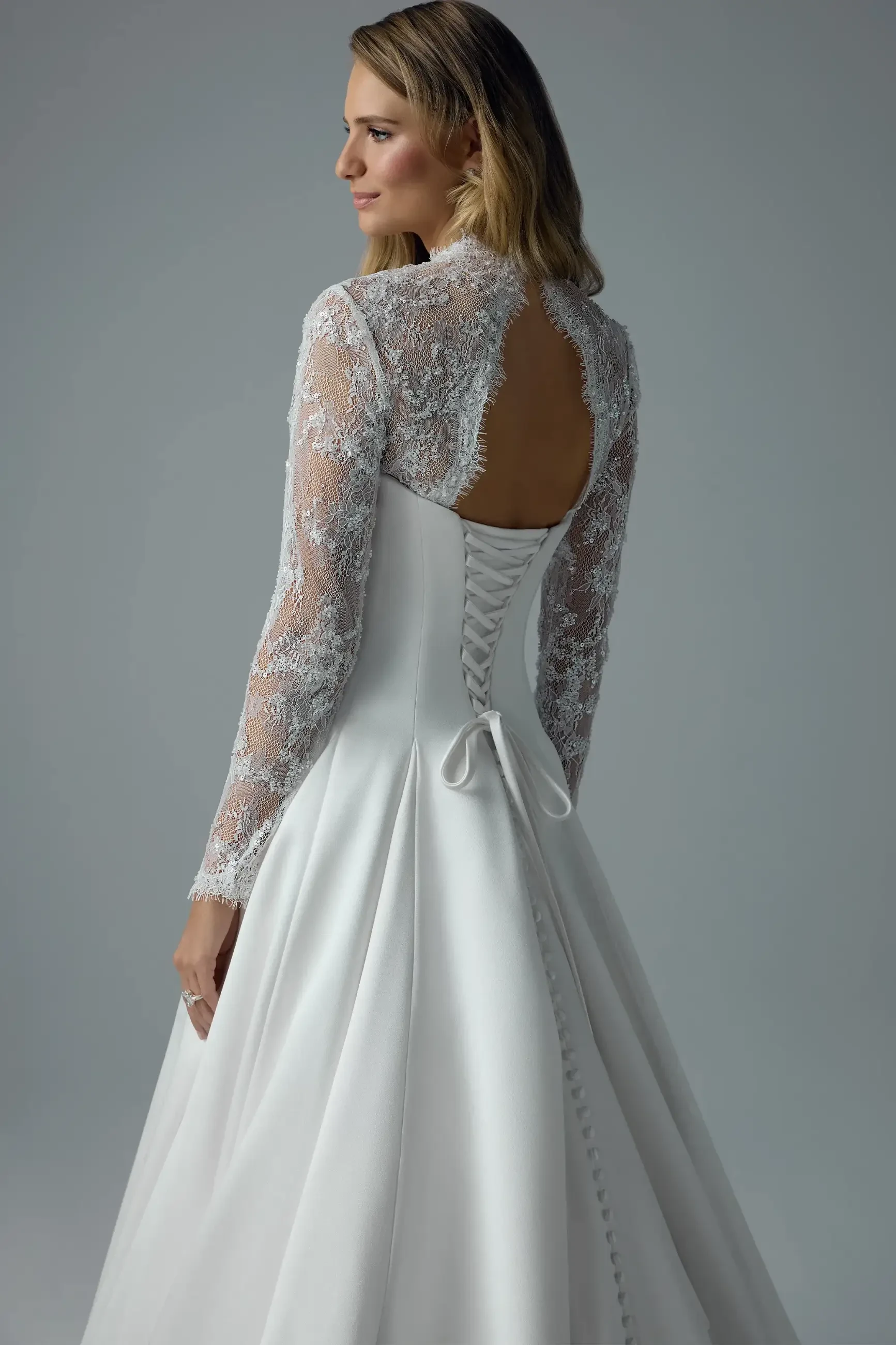 A woman in a white wedding dress with lace long sleeves and a low back, featuring a bow at the waist and button details down the back.