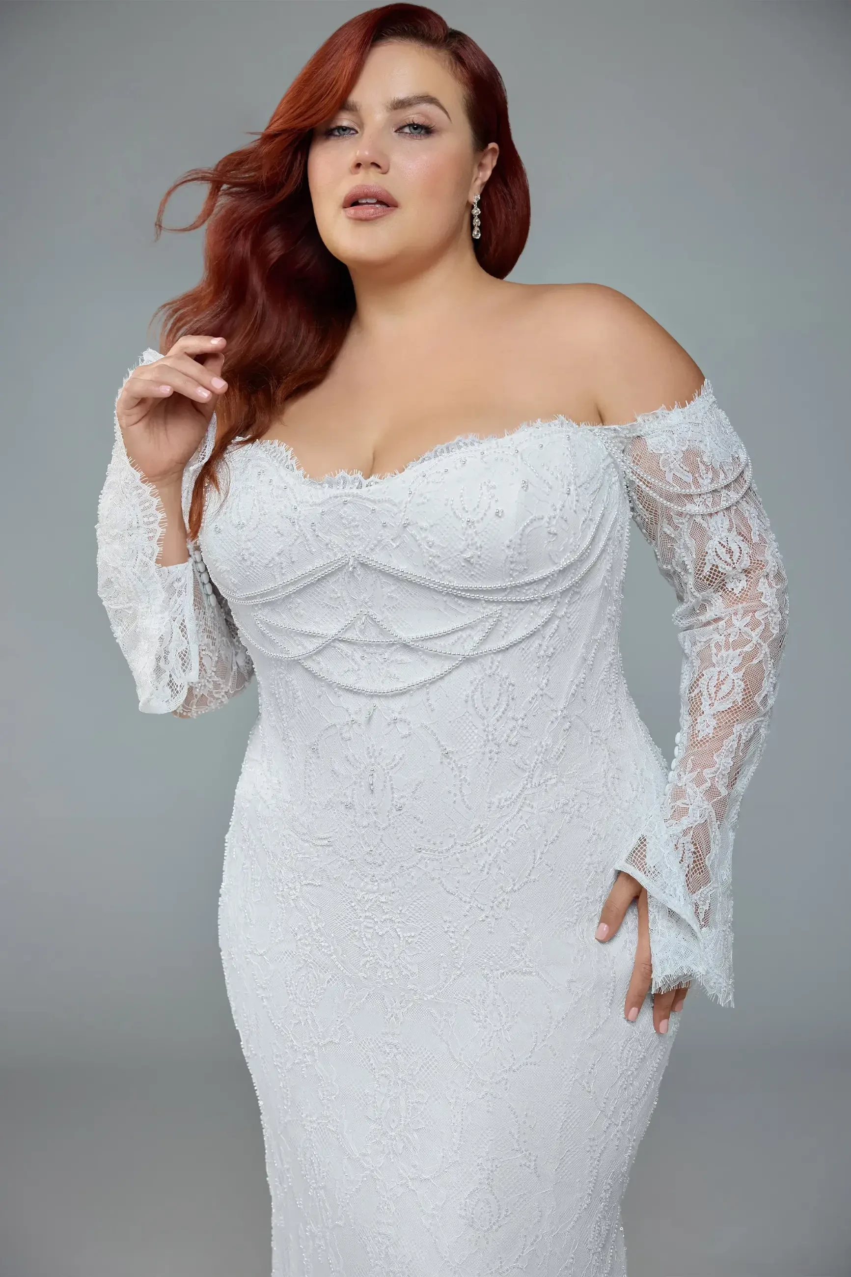 Woman in a white, off-shoulder lace wedding dress with long sleeves, looking confident. Her red hair cascades over her shoulder. Neutral background.