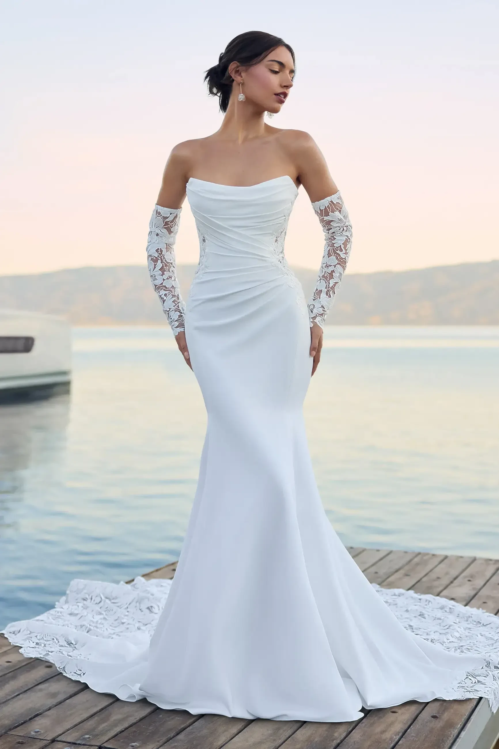 Model is wearing a lightweight crepe gown with an off-shoulder long-sleeve design, soft scoop neckline, floral lace bodice, and a dramatic laser-cut illusion back train.