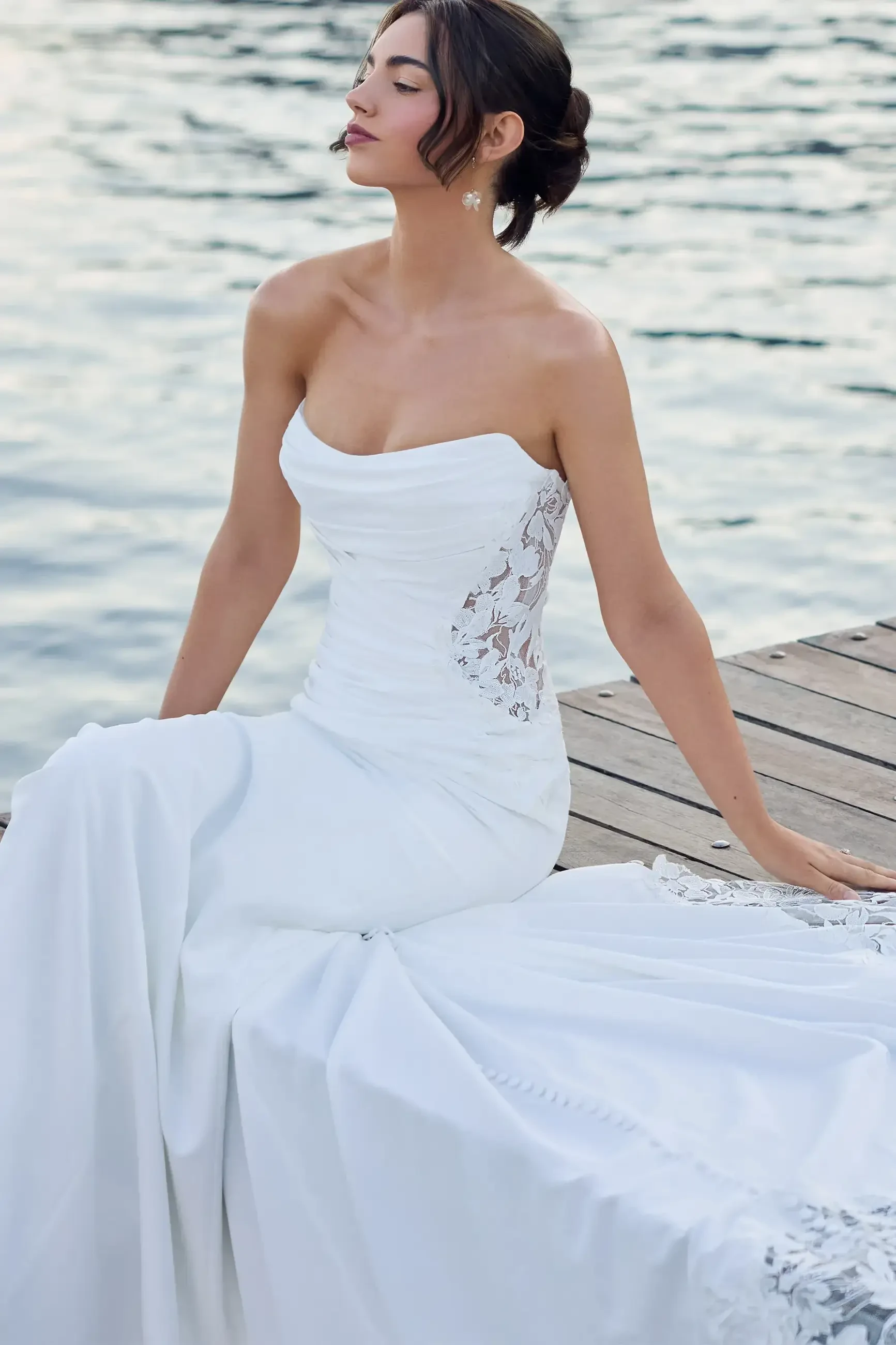 A woman sitting on a wooden dock wearing a strapless white wedding dress, with elegant lace detailing, against a backdrop of water.
