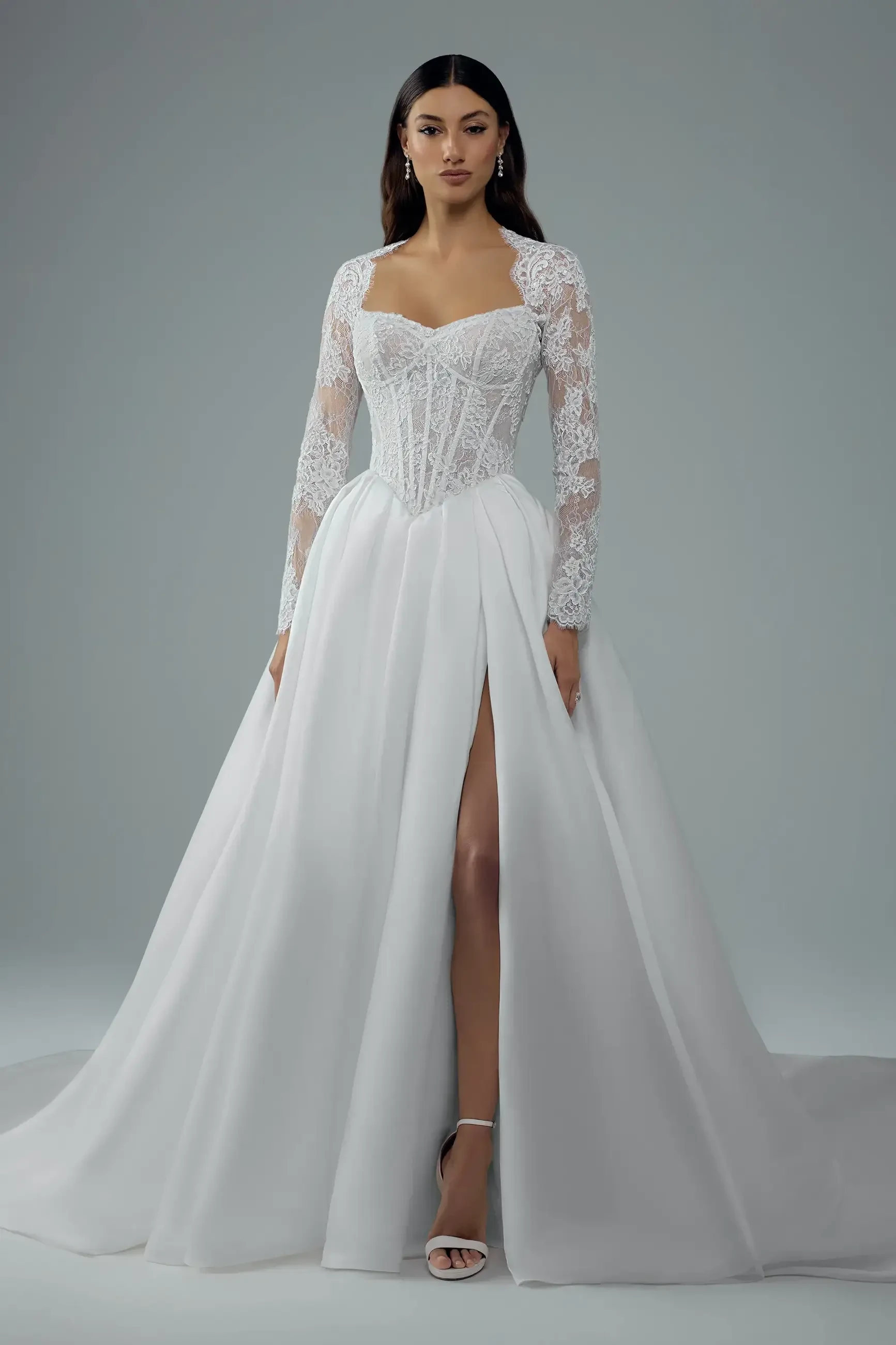 A model wearing an elegant white gown with long lace sleeves and a high slit. The dress features a fitted bodice and flows into a full skirt.
