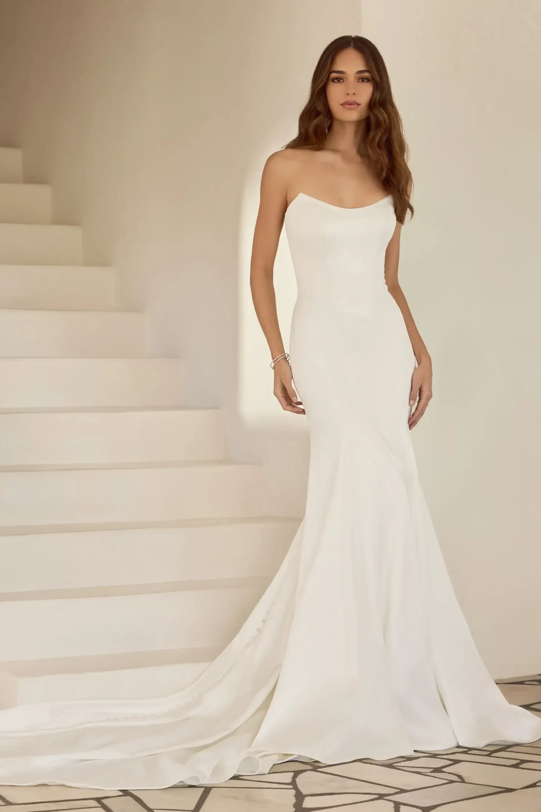 A model wearing a sleek, white strapless gown with a train, standing on a staircase.