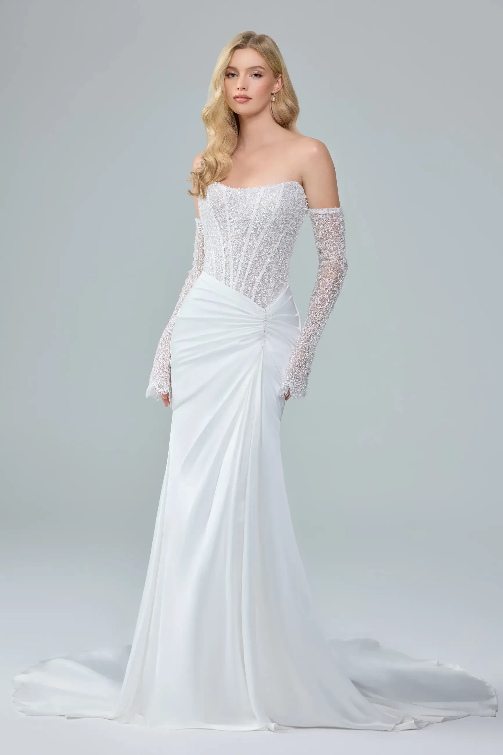 A model in an elegant white off-the-shoulder wedding dress with long lace sleeves, standing against a light gray background.