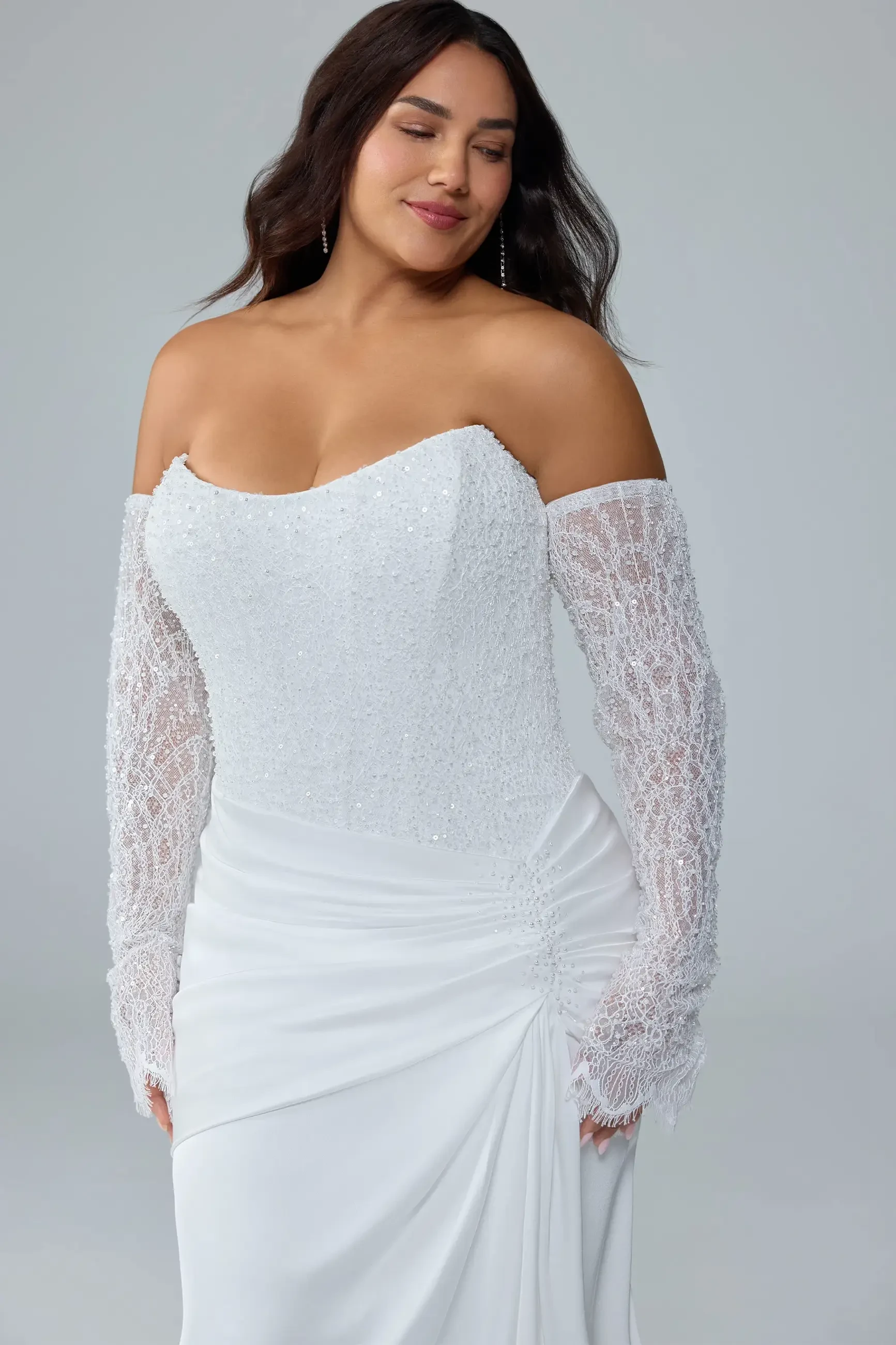 A woman wearing a white off-the-shoulder dress with intricate lace long sleeves, smiling softly against a neutral background.