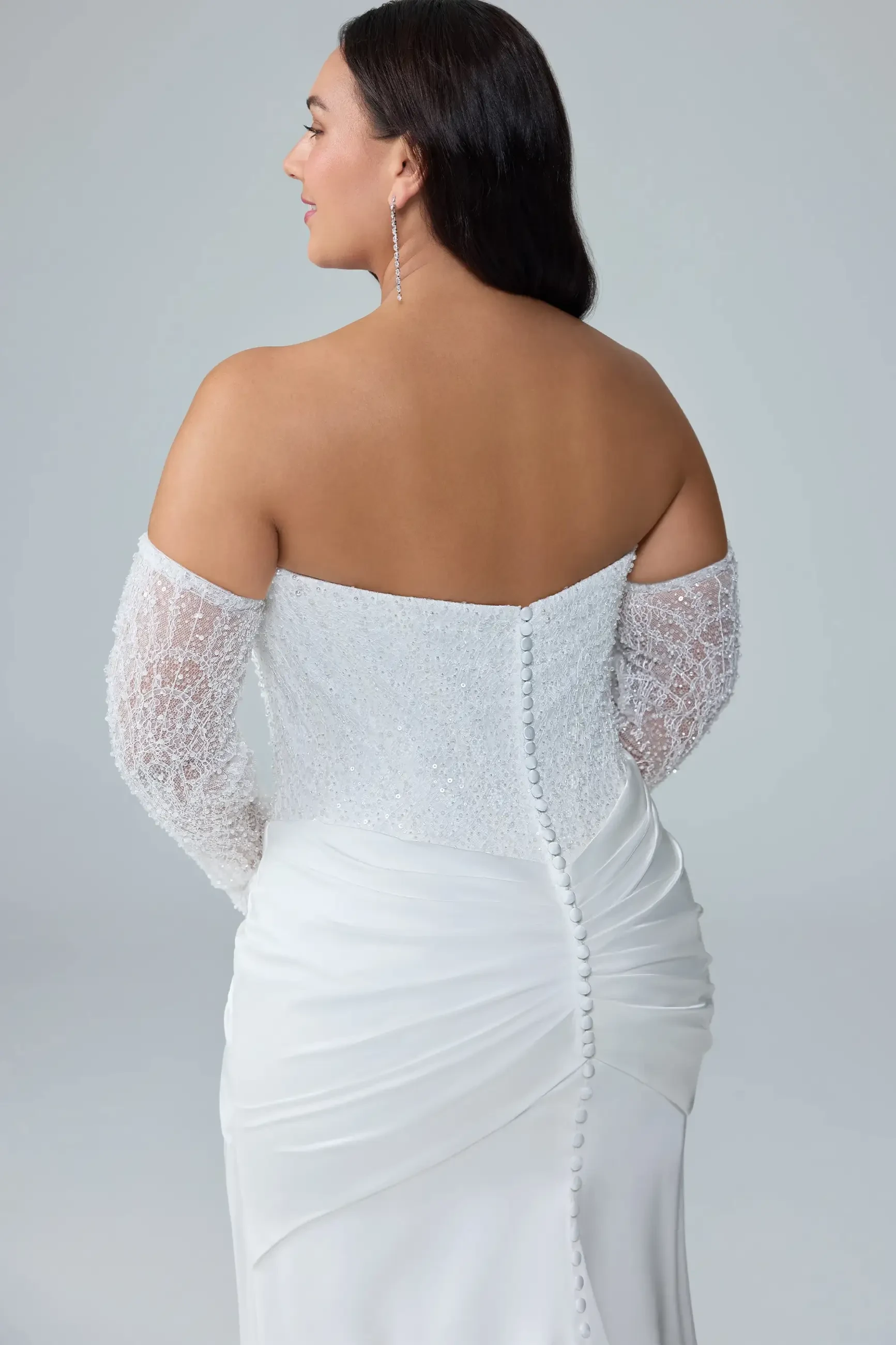 A woman wearing a white off-shoulder dress with long, beaded sleeves, seen from the back. The dress features a fitted silhouette with gathered fabric at the waist and buttons running down the back.
