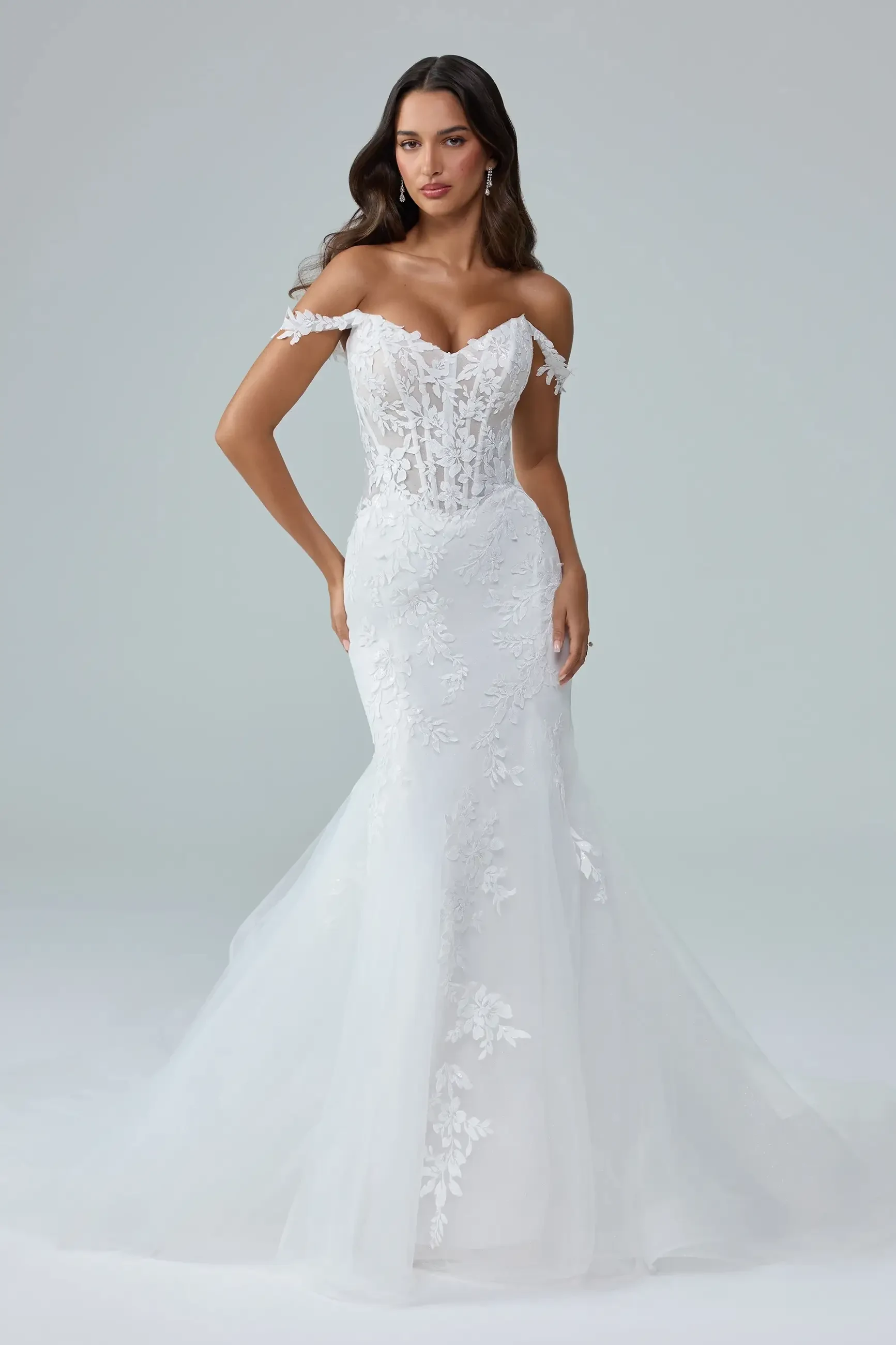 A woman wearing a beautiful white wedding dress with off-the-shoulder sleeves, a fitted bodice, and intricate lace detailing.