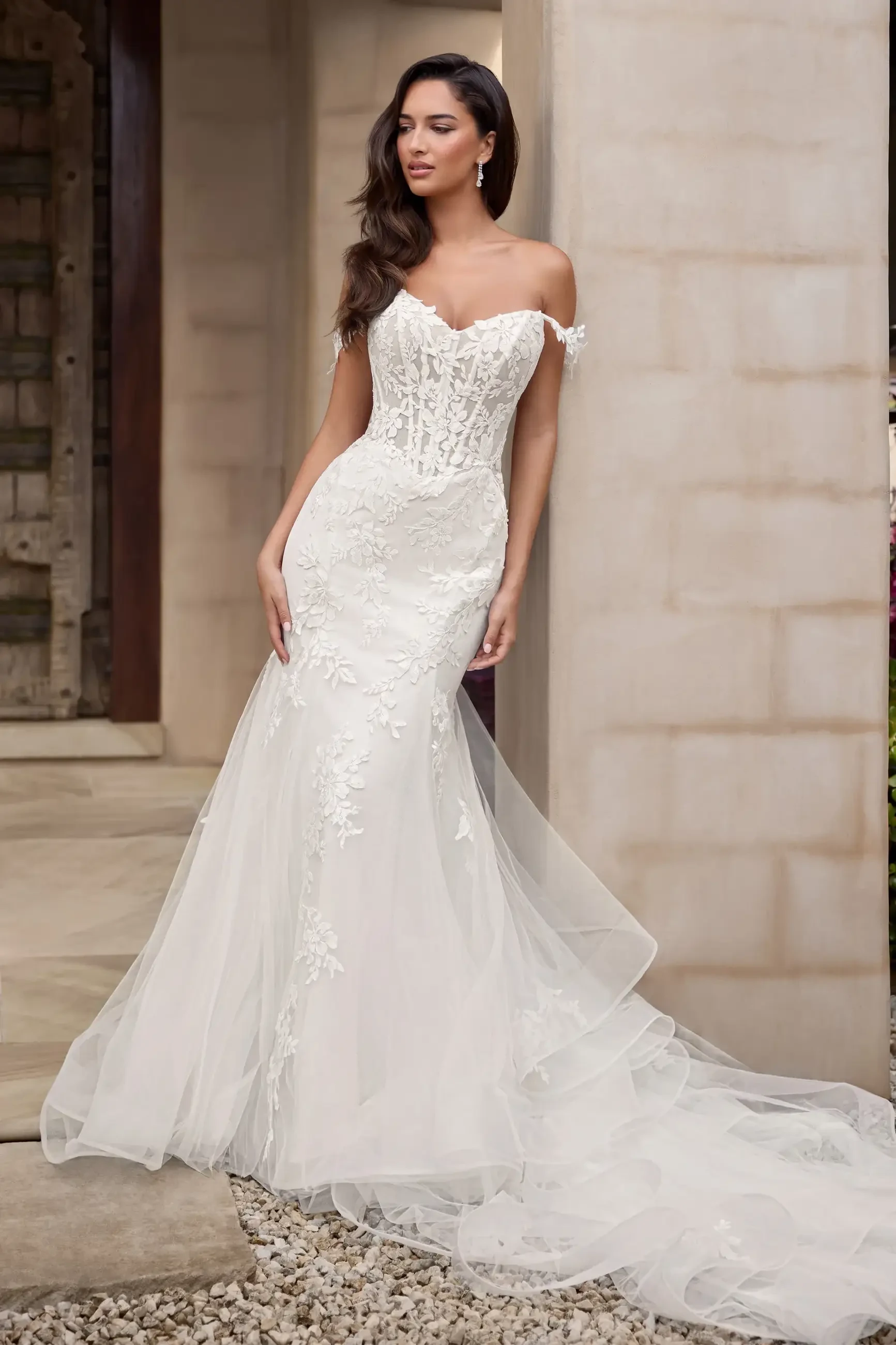 A bride in an elegant off-shoulder wedding dress with intricate lace and tulle details, posed beautifully outdoors.