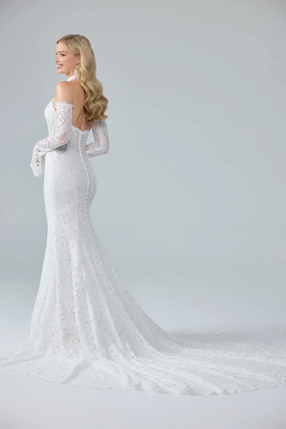 A model wearing a form-fitting white lace gown with off-the-shoulder long sleeves, standing against a light gray background.
